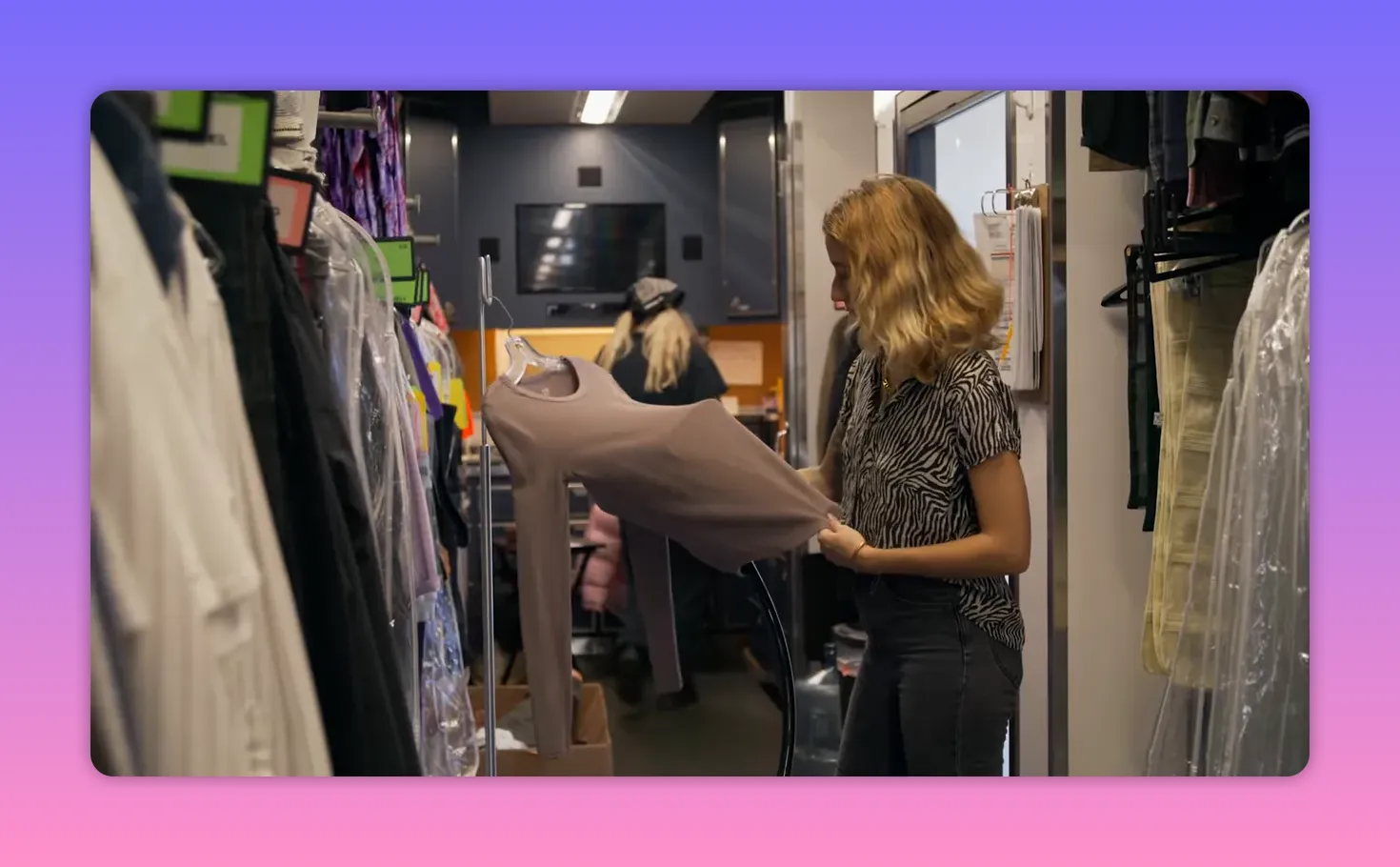 On-set costumer holding a shirt on a hanger inside a wardrobe truck, preparing and steaming a garment with organized racks in the background.