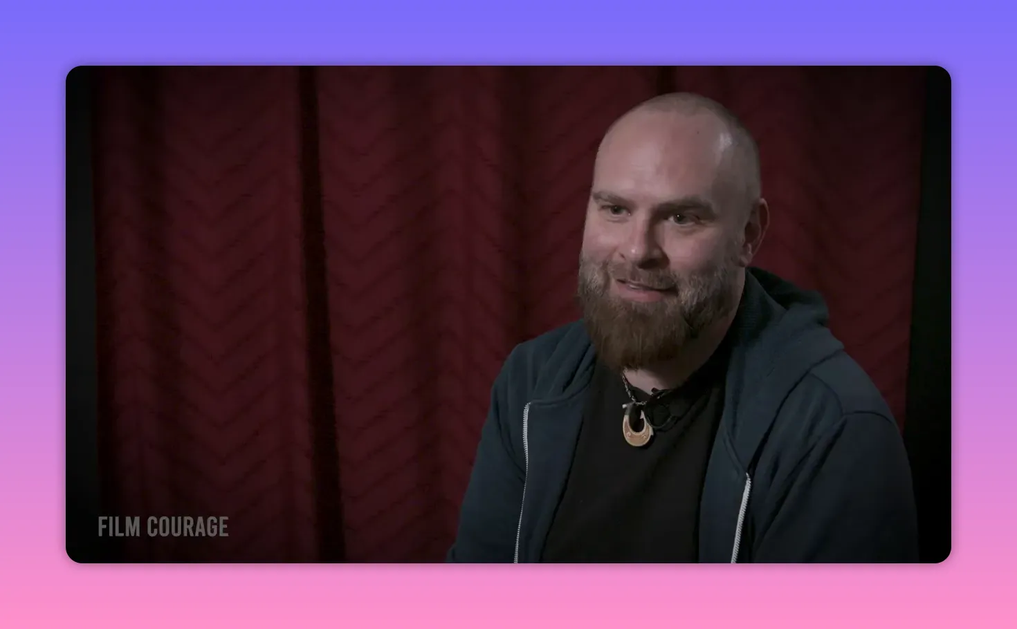 Interview subject smiling warmly and speaking, well-lit against a red curtain backdrop.