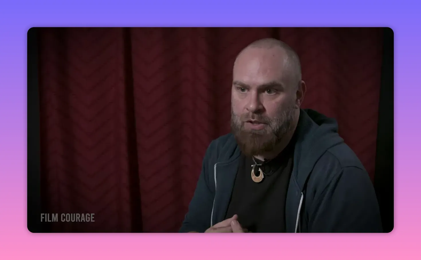 Well‑lit interview portrait of a bearded man speaking with hands clasped, red curtain backdrop.