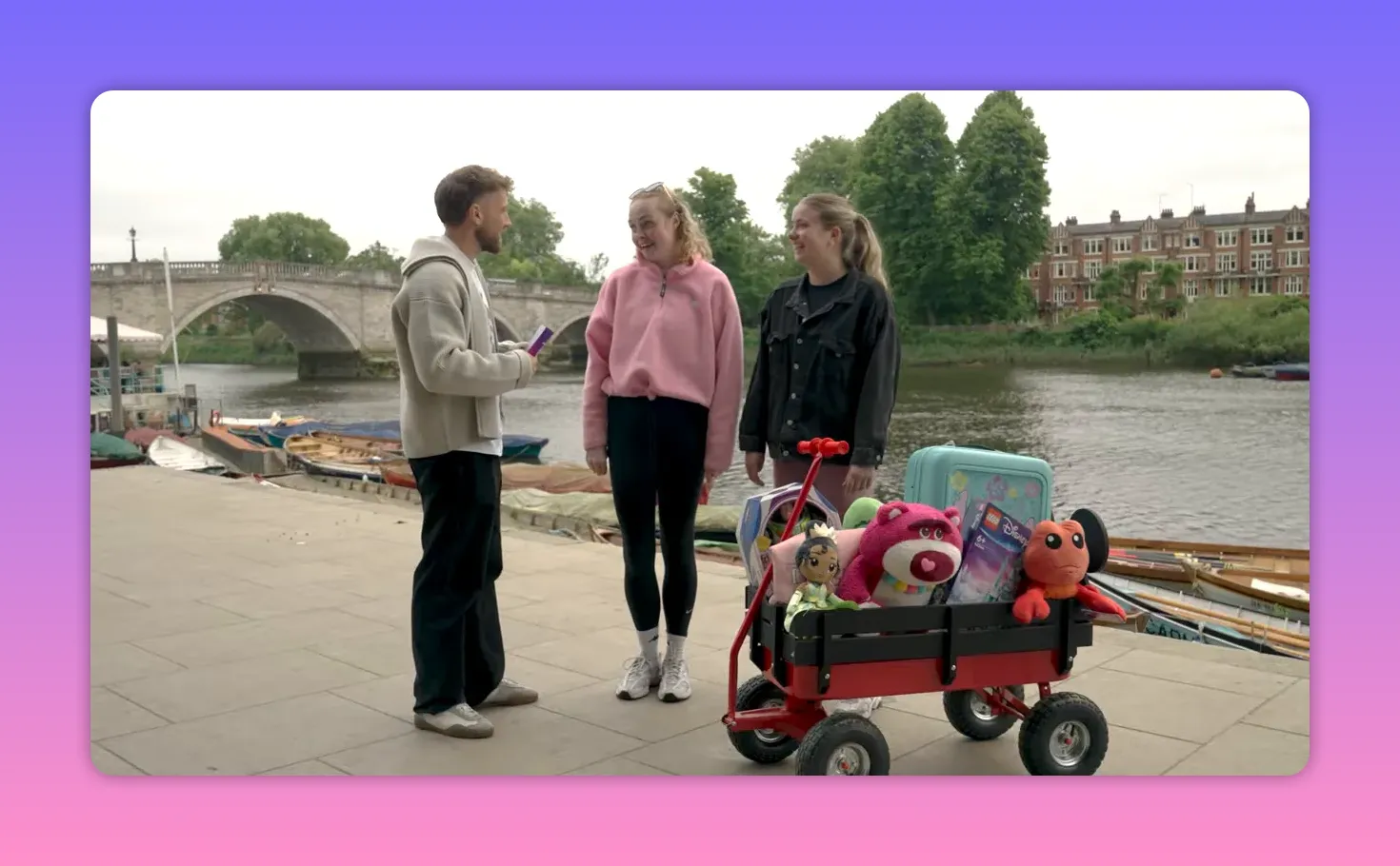 Host talking to two contestants standing next to a small red wagon packed with plush toys and Disney-themed prizes on a riverside walkway