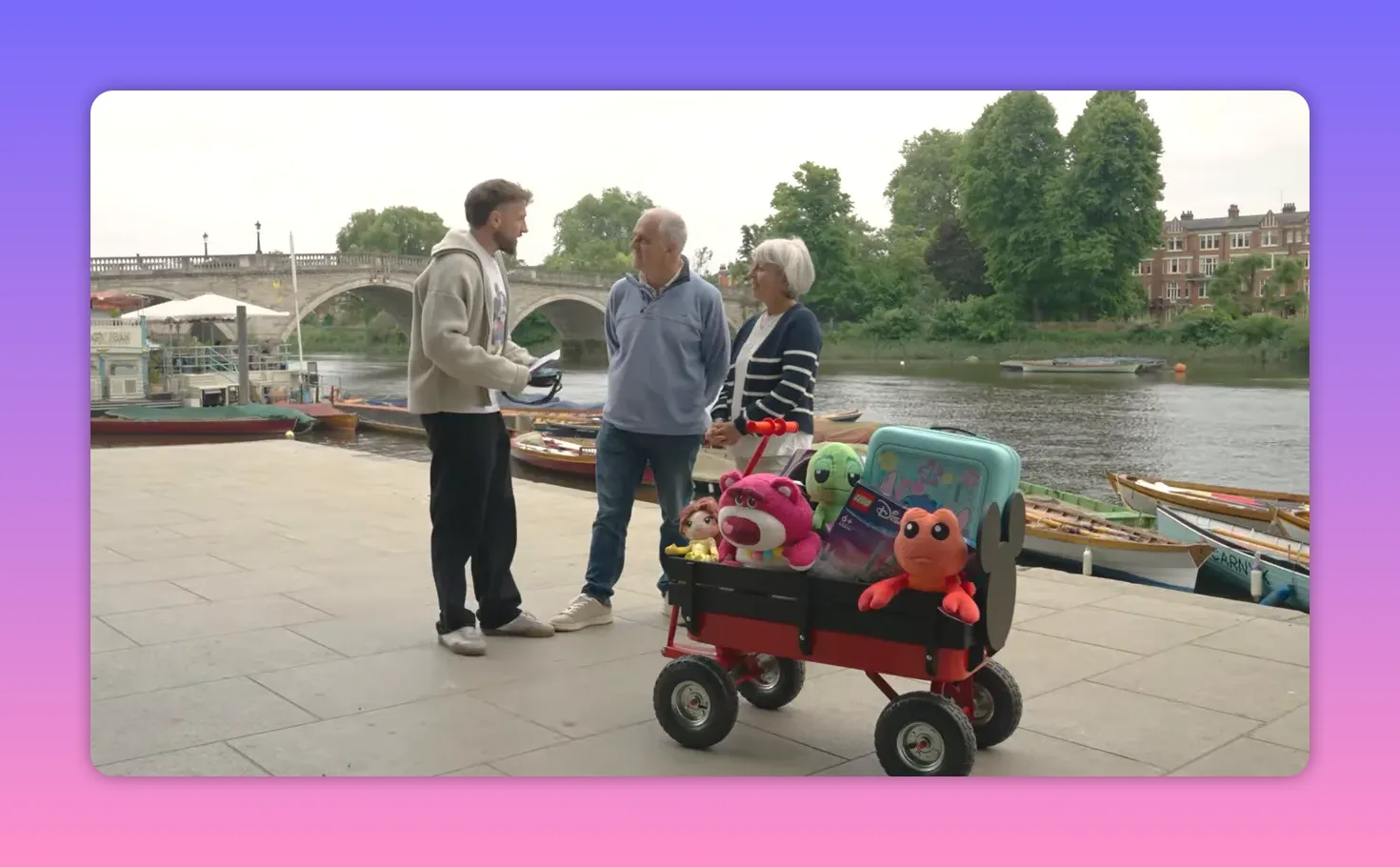 Wide clear shot of the host speaking with an older couple next to a small red wagon full of Disney-themed plush toys and prizes on a riverside walkway.