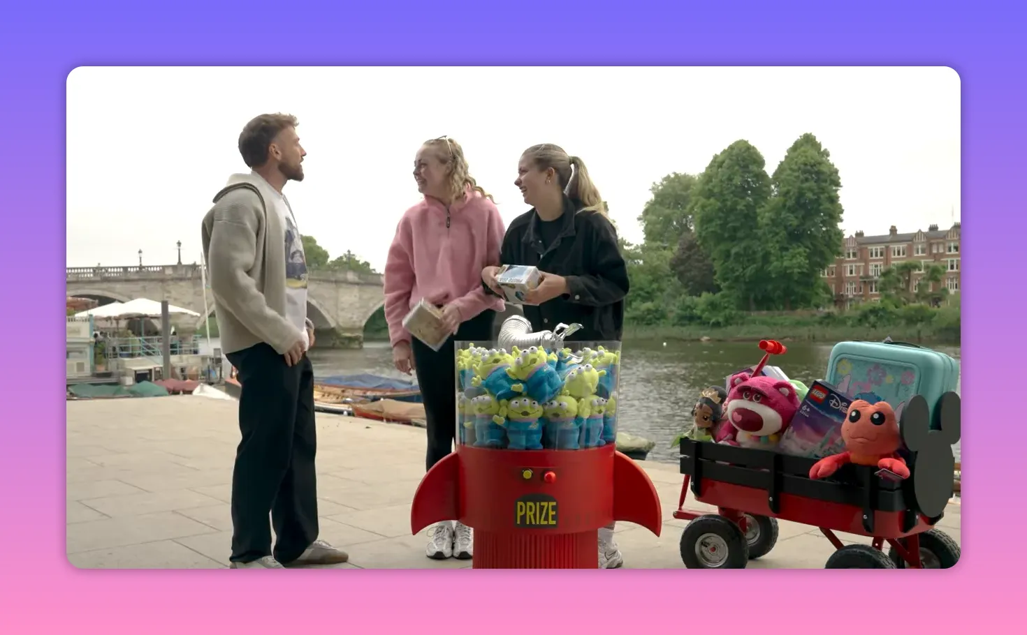 Host speaking with two contestants beside a riverside claw prize machine and a small red wagon full of Disney-themed toys and collectibles