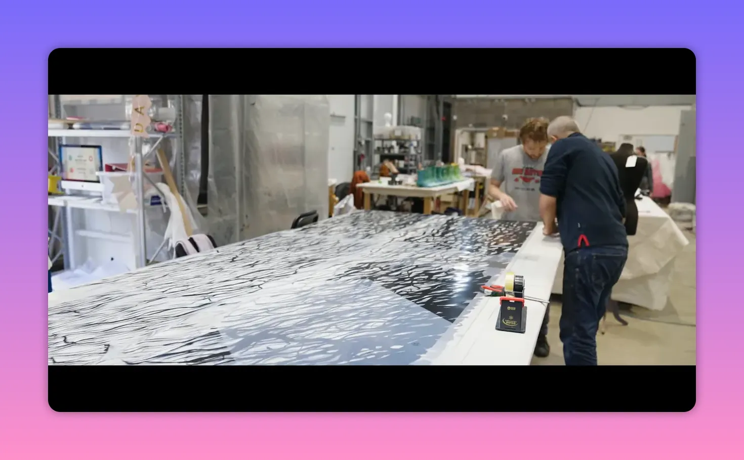 Wide shot of a long work table covered with printed fabric and two people working together in a textile workshop