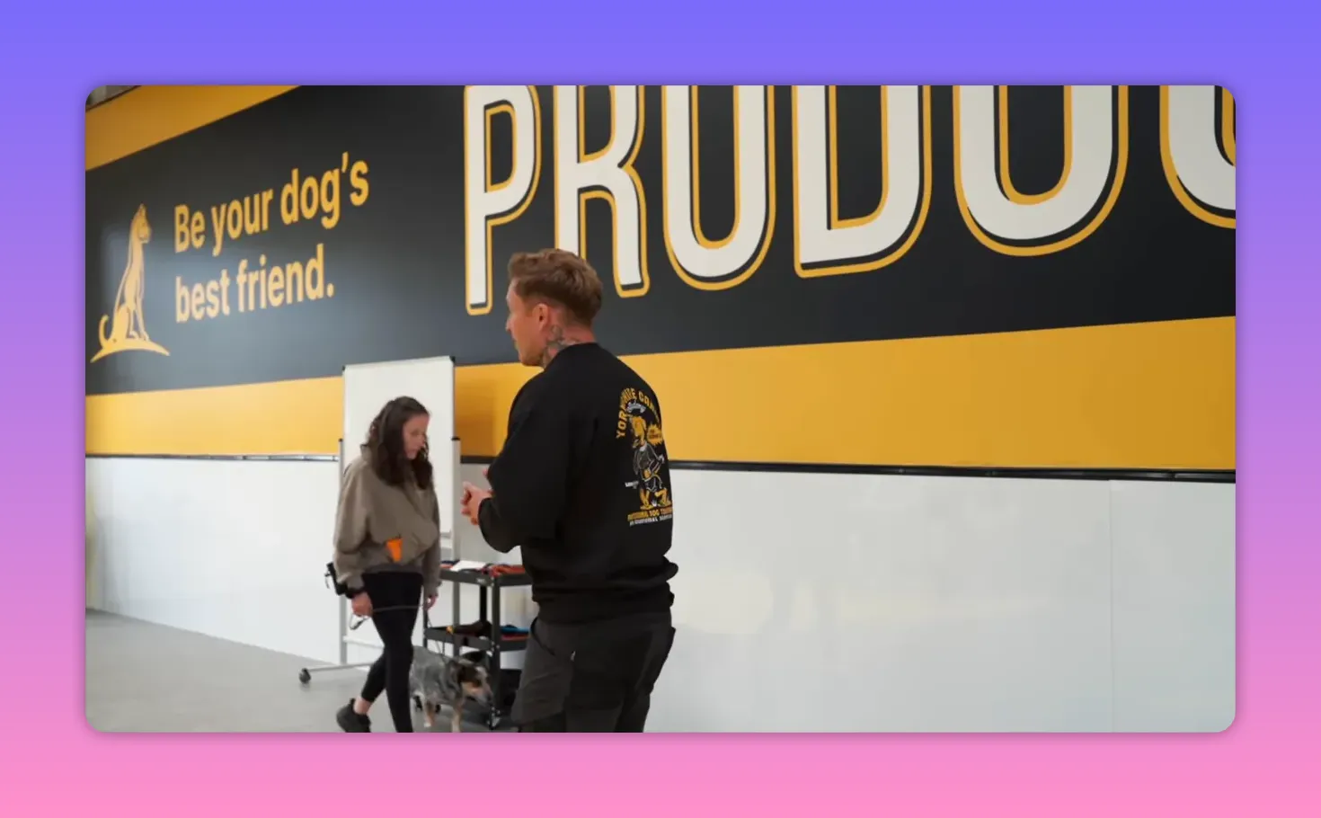 trainer coaching an owner who is walking a dog in an indoor training class in front of a bold Proudog mural