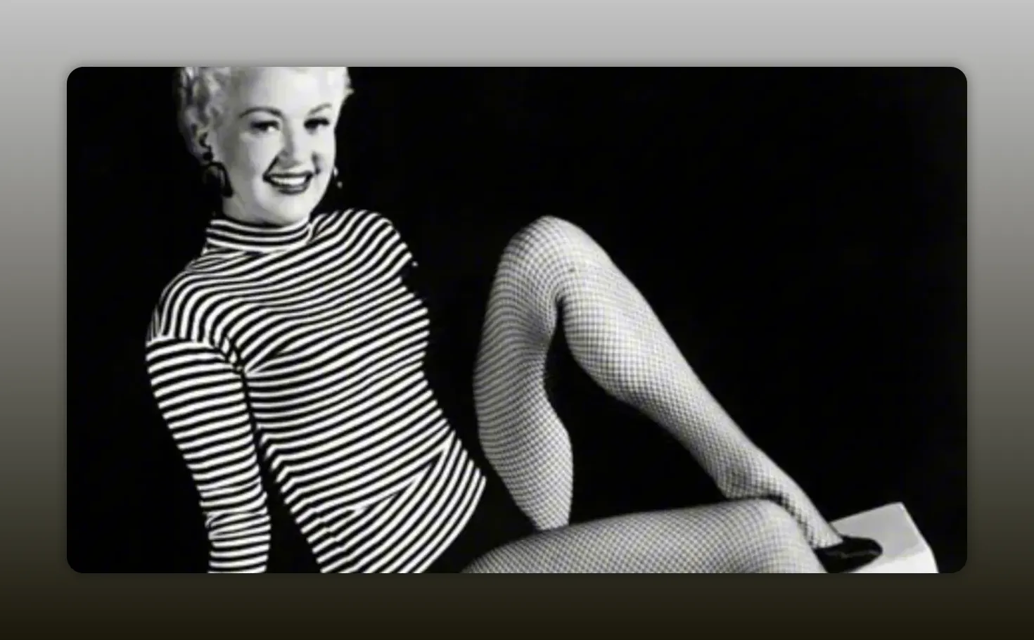 Sharper black-and-white promotional portrait of a smiling female performer in a striped shirt and fishnet stockings, seated with one leg raised and heels visible.