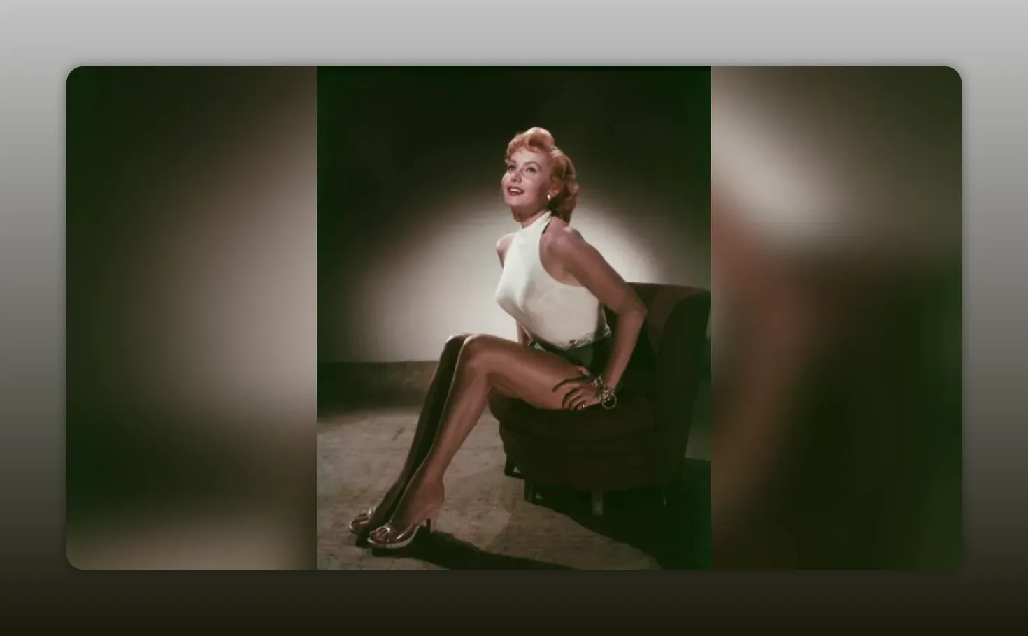 Clear vintage portrait of Betty Grable seated showing her legs in a white top and heels, full-body studio shot