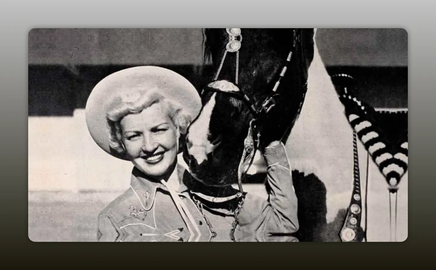 Vintage black-and-white image of a smiling woman in a cowgirl outfit holding a horse's head close, clear detail of bridle and costume.