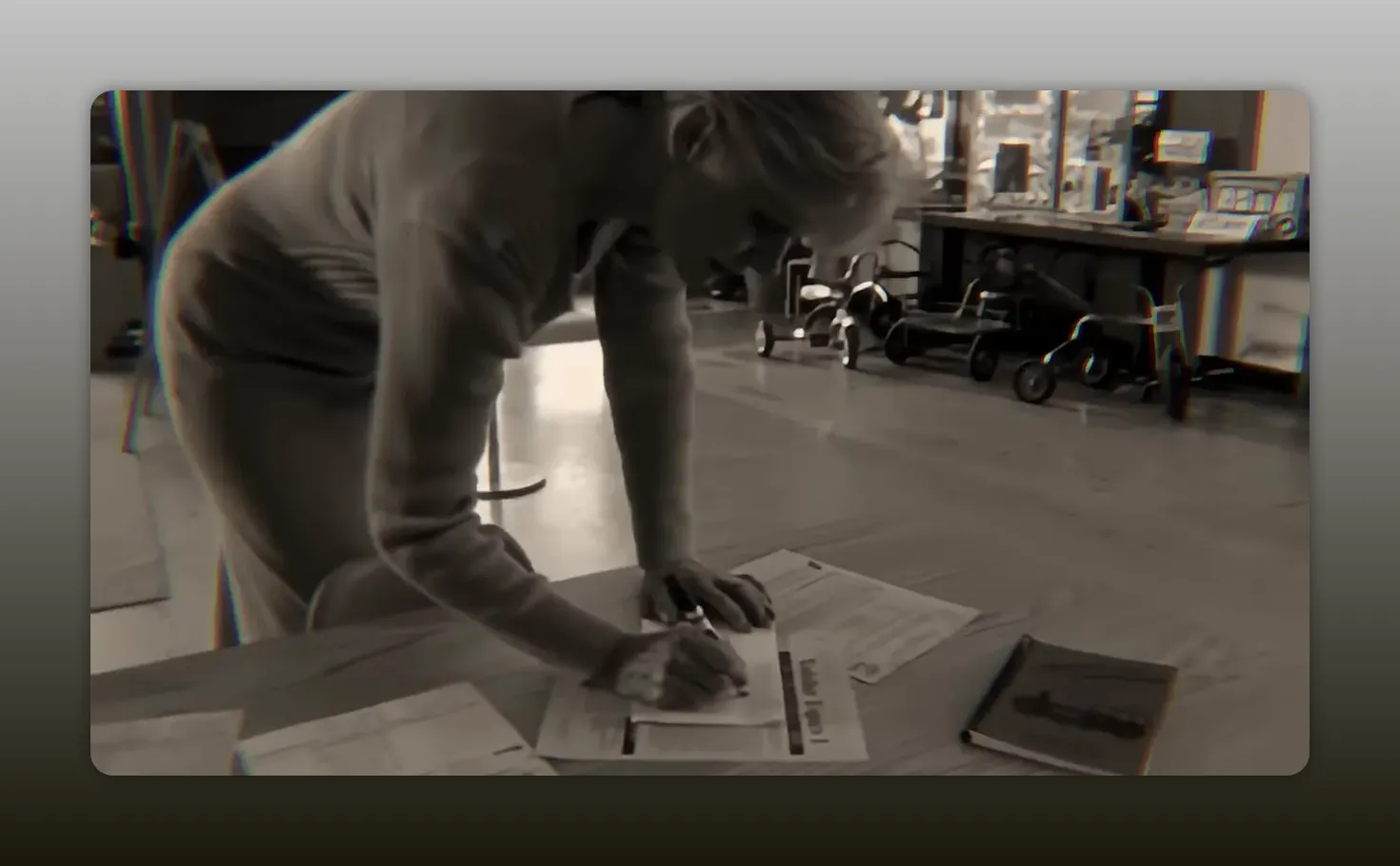 Preschool teacher leaning over a table writing on a paper in a lobby with children's tricycles behind her