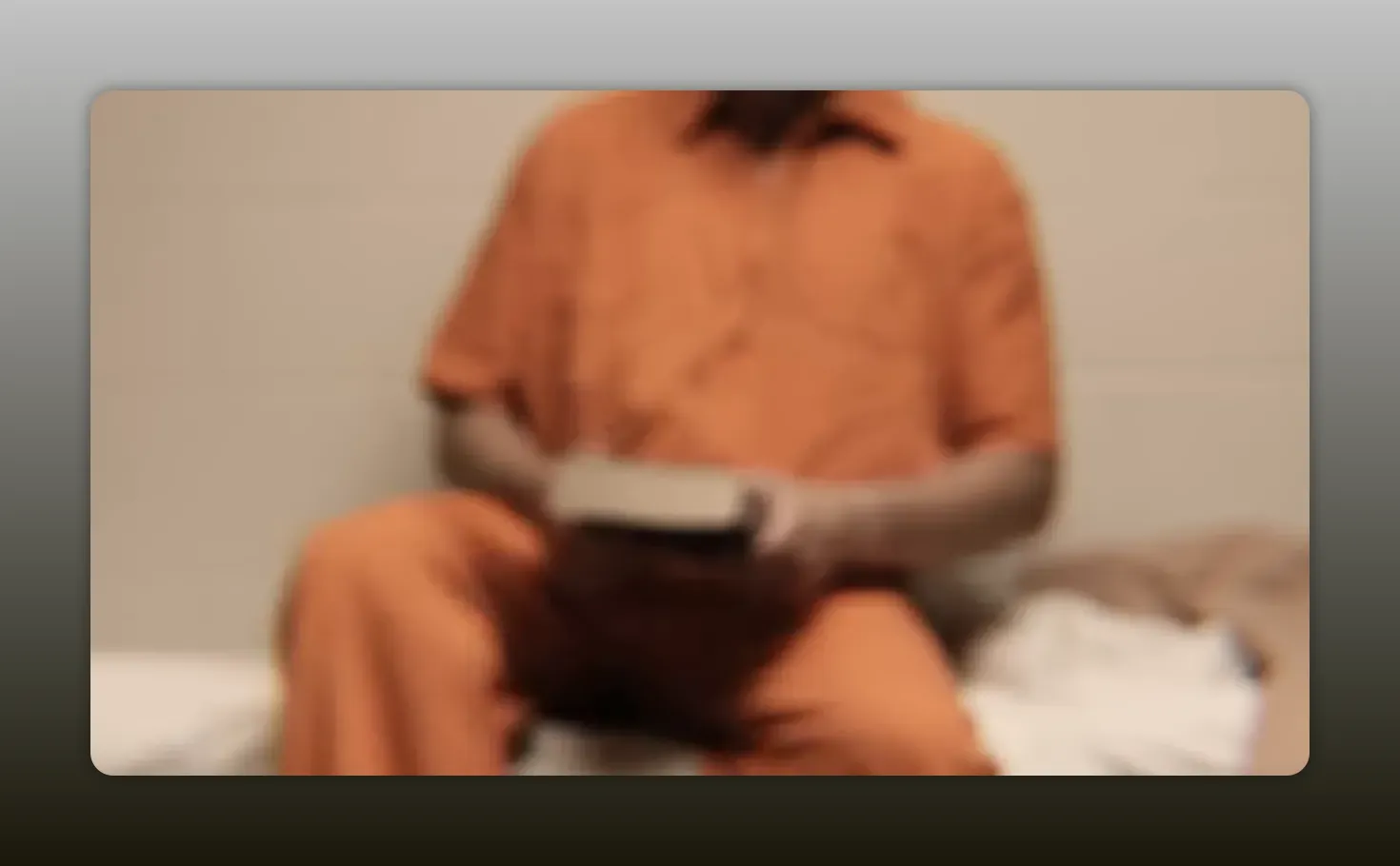 Out-of-focus seated figure in an orange prison jumpsuit, hands holding an object