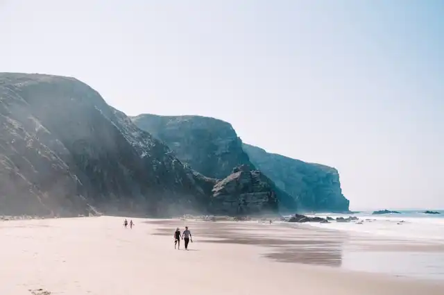 Scenic beach with towering cliffs and people walking along the shore under a clear blue sky in Portugal.