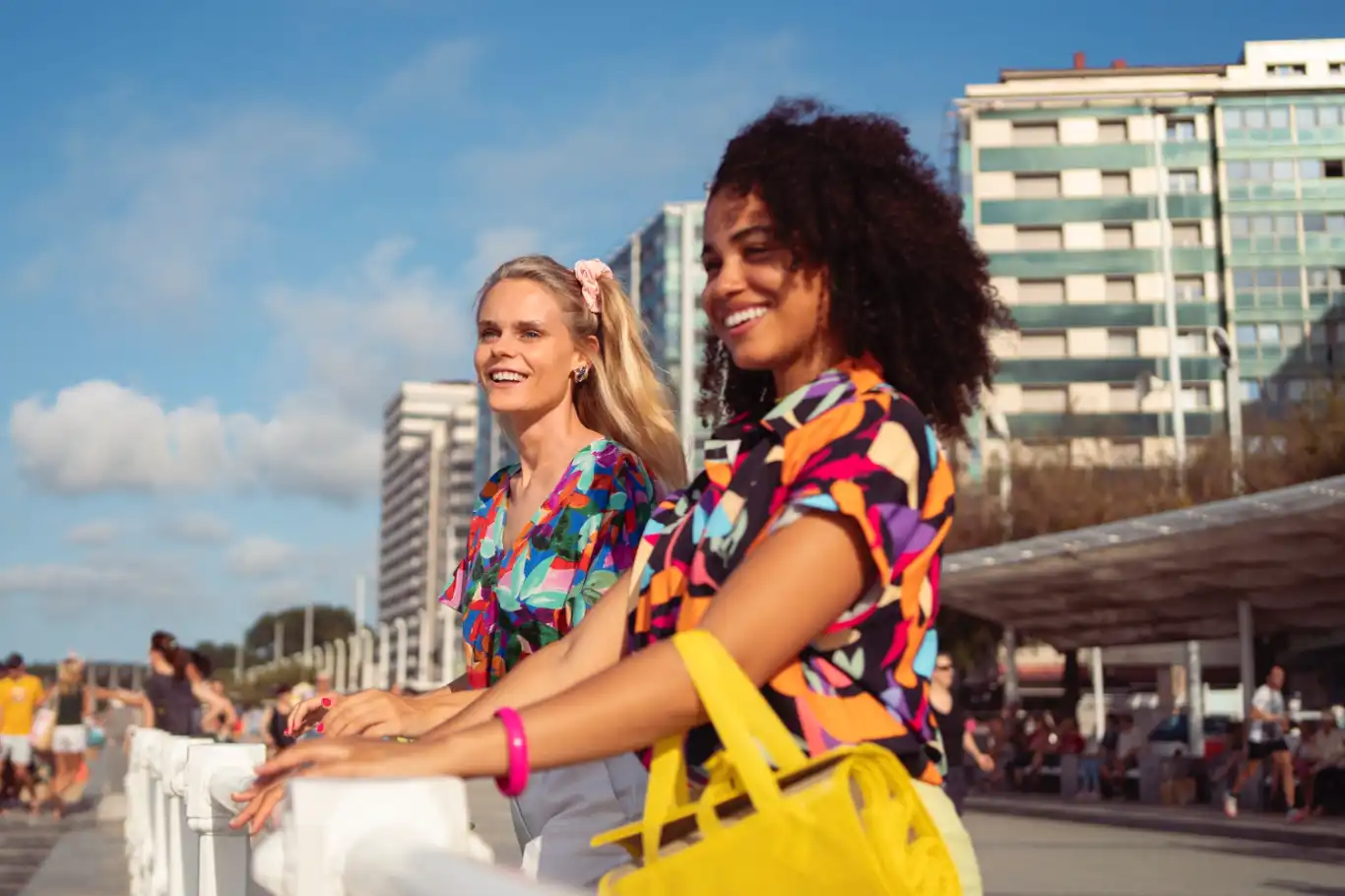Two women enjoying the sunny day at a promenade in Rio de Janeiro, smiling and looking out over the beach, with modern buildings in the background.