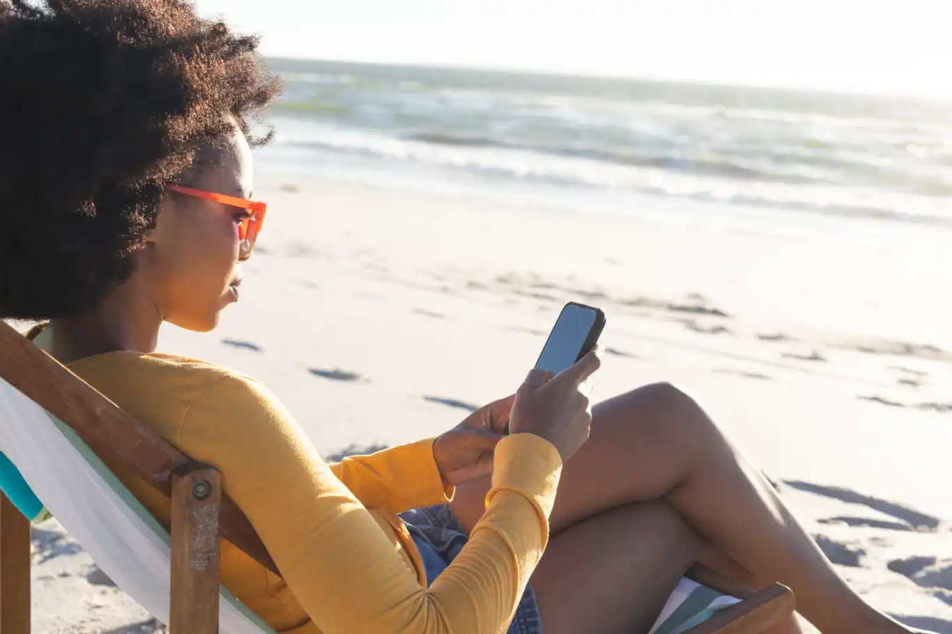 Woman in sunglasses sitting in deckchair using smartphone on sunny beach. Summer, relaxation, communication, free time and vacation, unaltered.