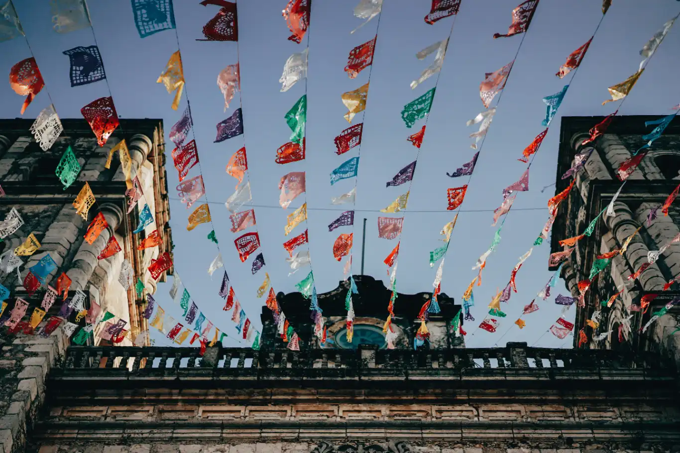 Colorful papel picado streamers fluttering in the breeze above a historical building's façade under a clear blue sky.