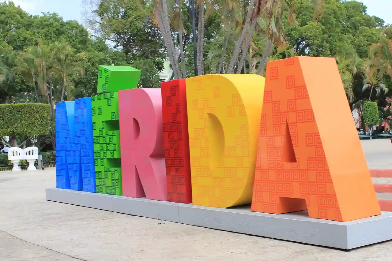 Colorful three-dimensional 'MÉRIDA' sign in bold letters, decorated with traditional patterns, standing in a sunny plaza with lush green trees in the background.