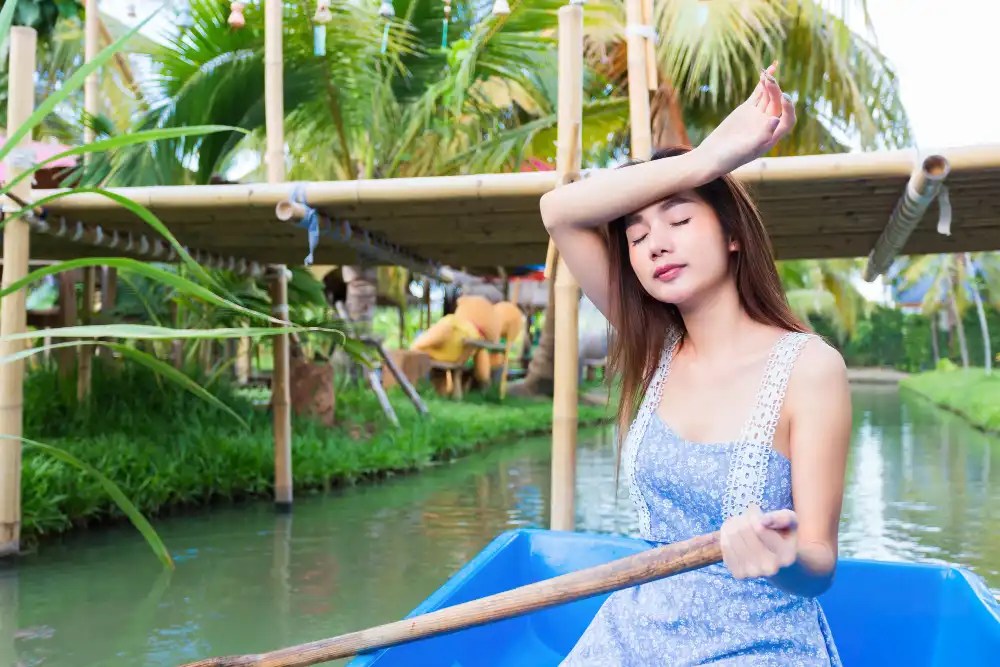 A woman in a blue dress paddling a boat and shielding her eyes from the sun, suggesting the heat, in a lush canal setting.