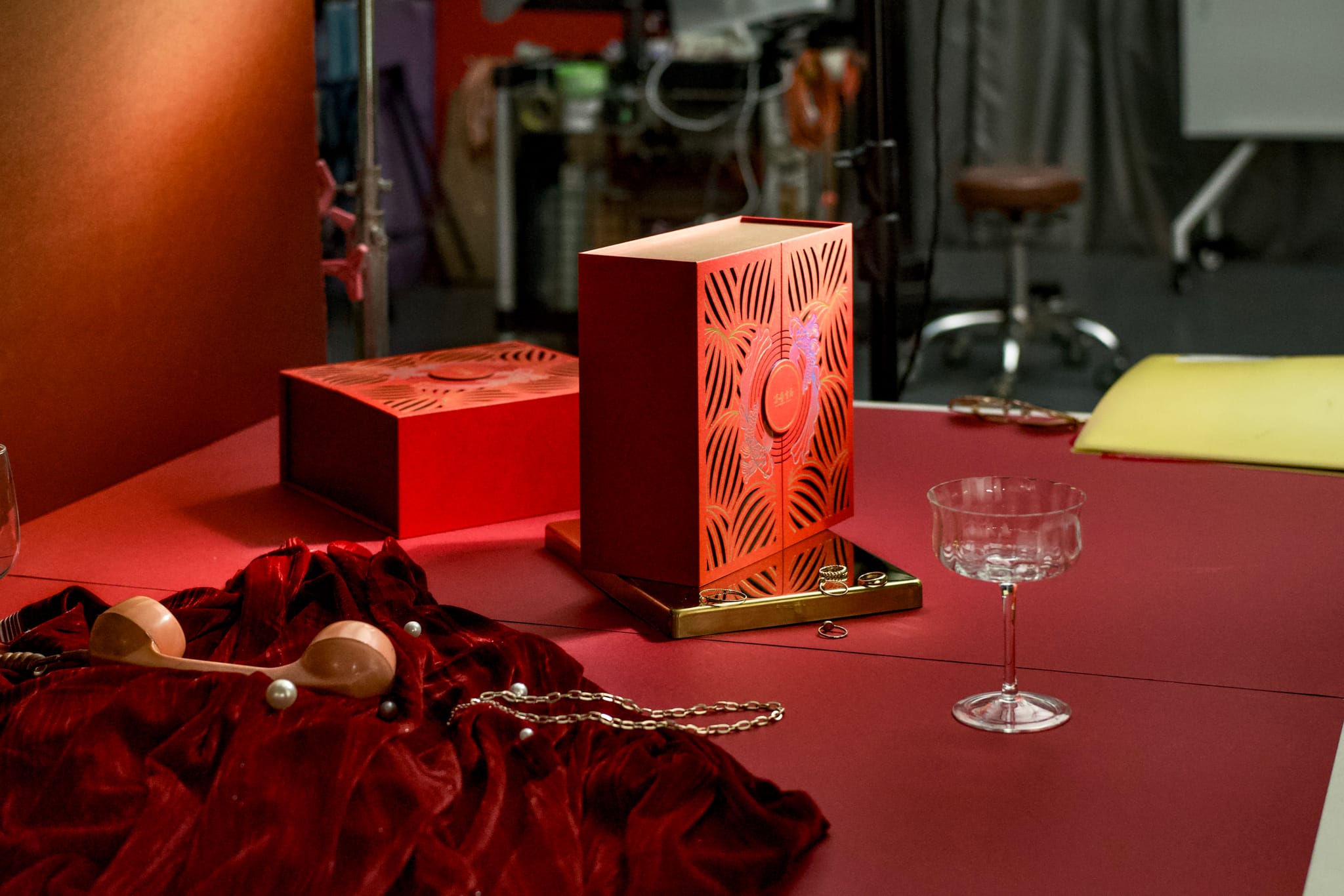攝影棚內景，紅色調背景上，禮盒、深紅絲絨與珠寶擺設，捕捉華麗慵懶的視覺氛圍。