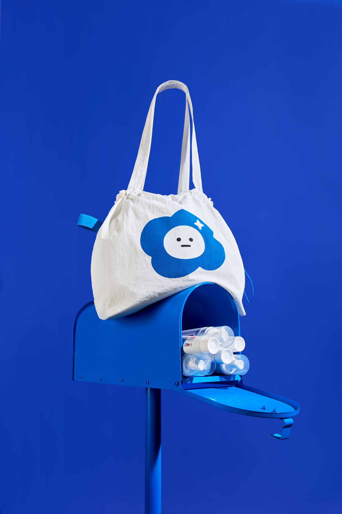 藍色背景下，一個藍色郵筒上放著印有AH!Q雲朵角色的白色帆布包，郵筒內露出聯名保養品。