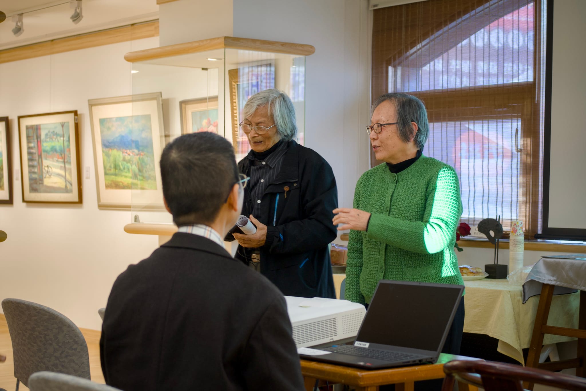 李季眉館長（右）在「一條線的故事」展覽會場與來賓交流，背景是展示的畫作。前方一位聽眾的背影，捕捉了講座後與觀眾互動的溫馨氛圍。