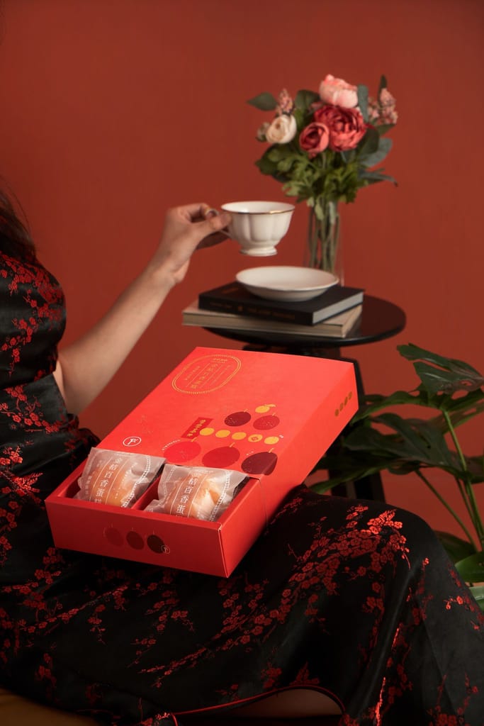 女子穿著典雅服飾，手持茶杯，膝上紅色新年禮盒內含點心，營造溫馨節慶感。