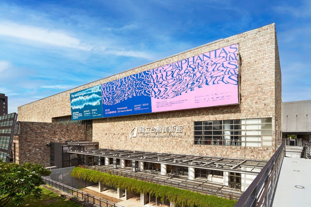 國立台灣美術館外觀，懸掛未至之城2021亞洲藝術雙聯展巨幅海報，藍天白雲下建築氣勢磅礴。