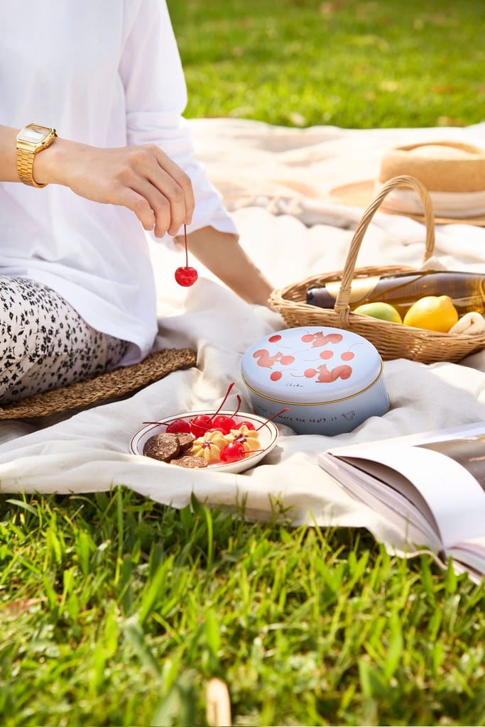 陽光草地上野餐情境，人物手持櫻桃，搭配品牌禮盒與精緻甜點。
