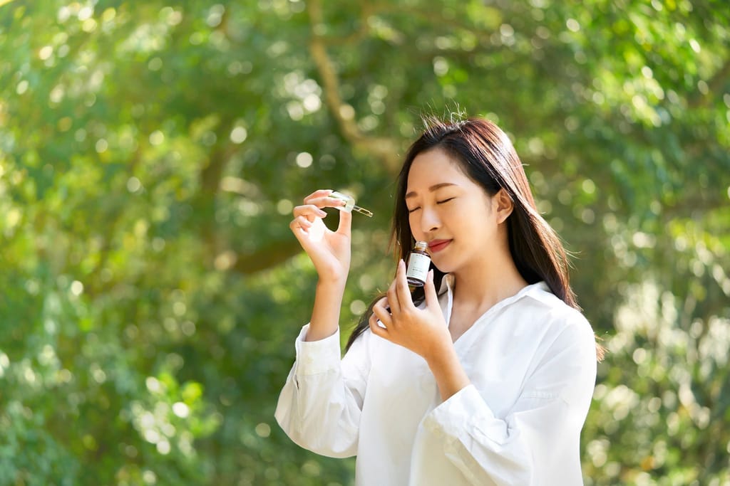 一位身穿白衣的女性在綠意盎然的戶外閉眼輕嗅保養品，感受來自大自然的純淨芳香。