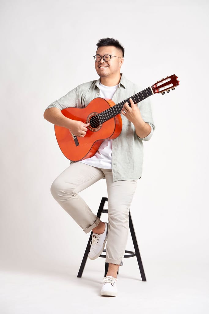 一位戴眼鏡的男性樂手坐著彈奏吉他，面帶微笑，展現真誠樣貌。