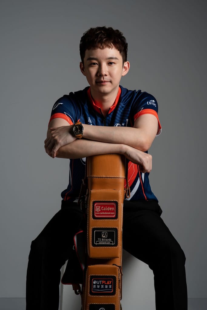 一位身著藍色運動服的年輕男性選手，雙臂交疊倚靠在專業棕色球桿箱上，目光堅定地凝視前方，展現專注且自信的形象。