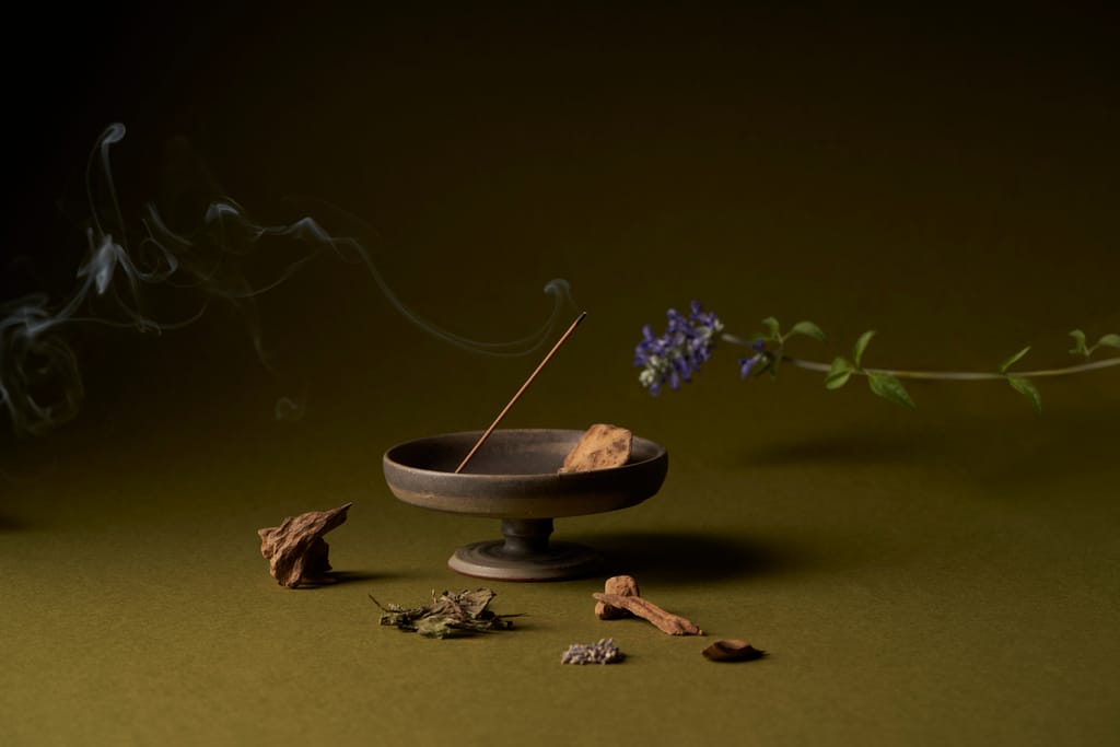 燃燒的天然線香在簡約香爐中，伴隨木塊、草本與花朵，煙霧裊裊，象徵著東方療癒與儀式感。