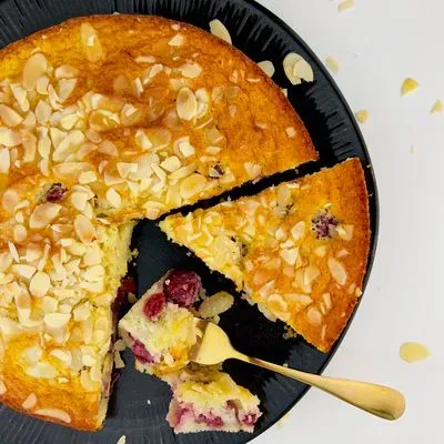  Un gâteau aux amandes parsemé d'éclats d'amandes, avec des griottes, servi sur une assiette noire avec une part entamée et une cuillère dorée.