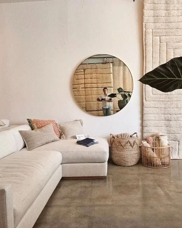 Living room corner with a beige sofa, decorative pillows, a round mirror on the wall reflecting a person, and baskets with textiles and a lamp.