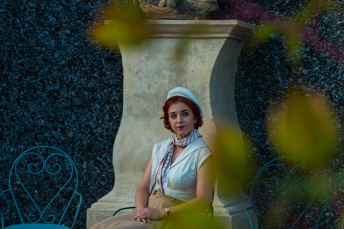 A red haired woman sits quietly in a courtyard, behind her a statue and a wall made of black pebbles. It looks like the photo was taken without her knowing she was being watched