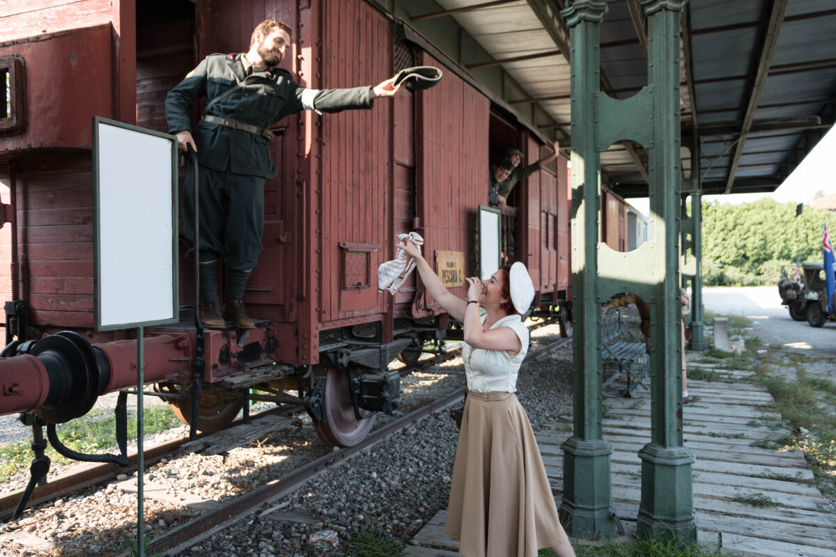 A soldier standing on a train waves his hat to a woman that's waving a tissue and weeping for his departure
