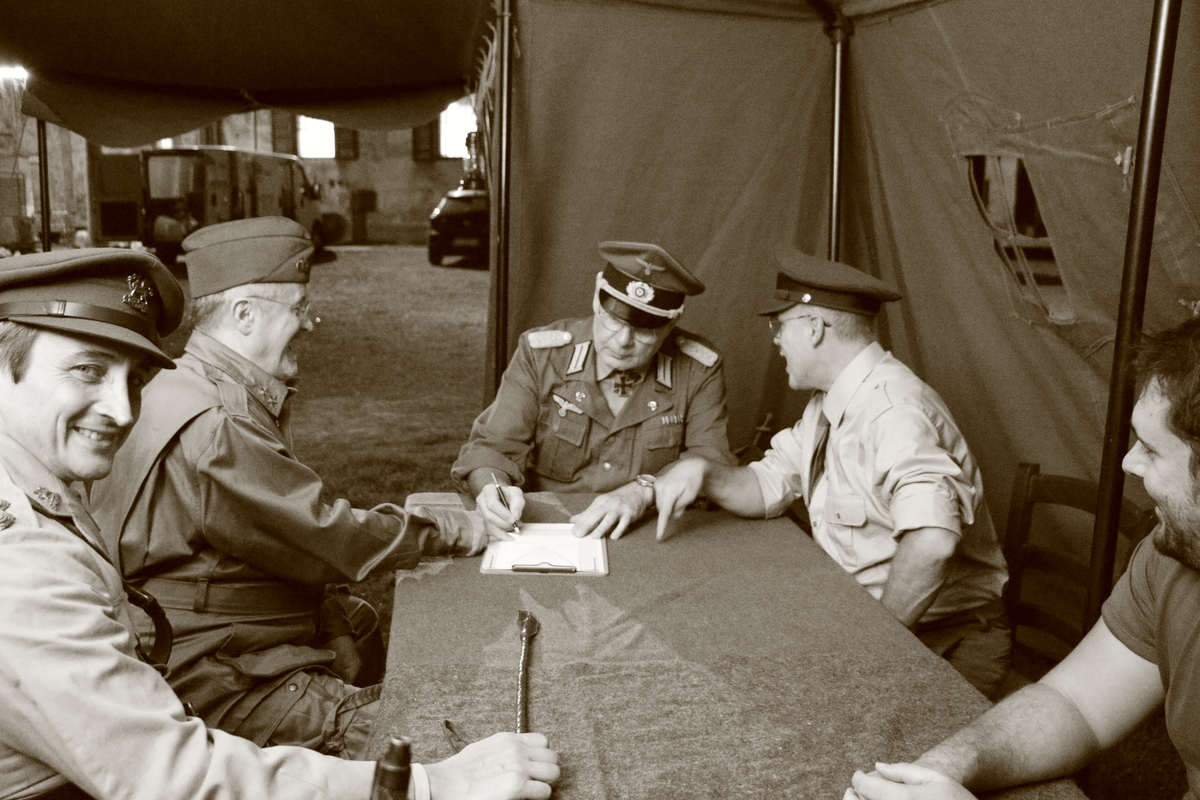 Reenactment, five people dressed as WW2 soldiers gather around a table, one of them dressed as a general is signing a piece of paper, while another points to the table.