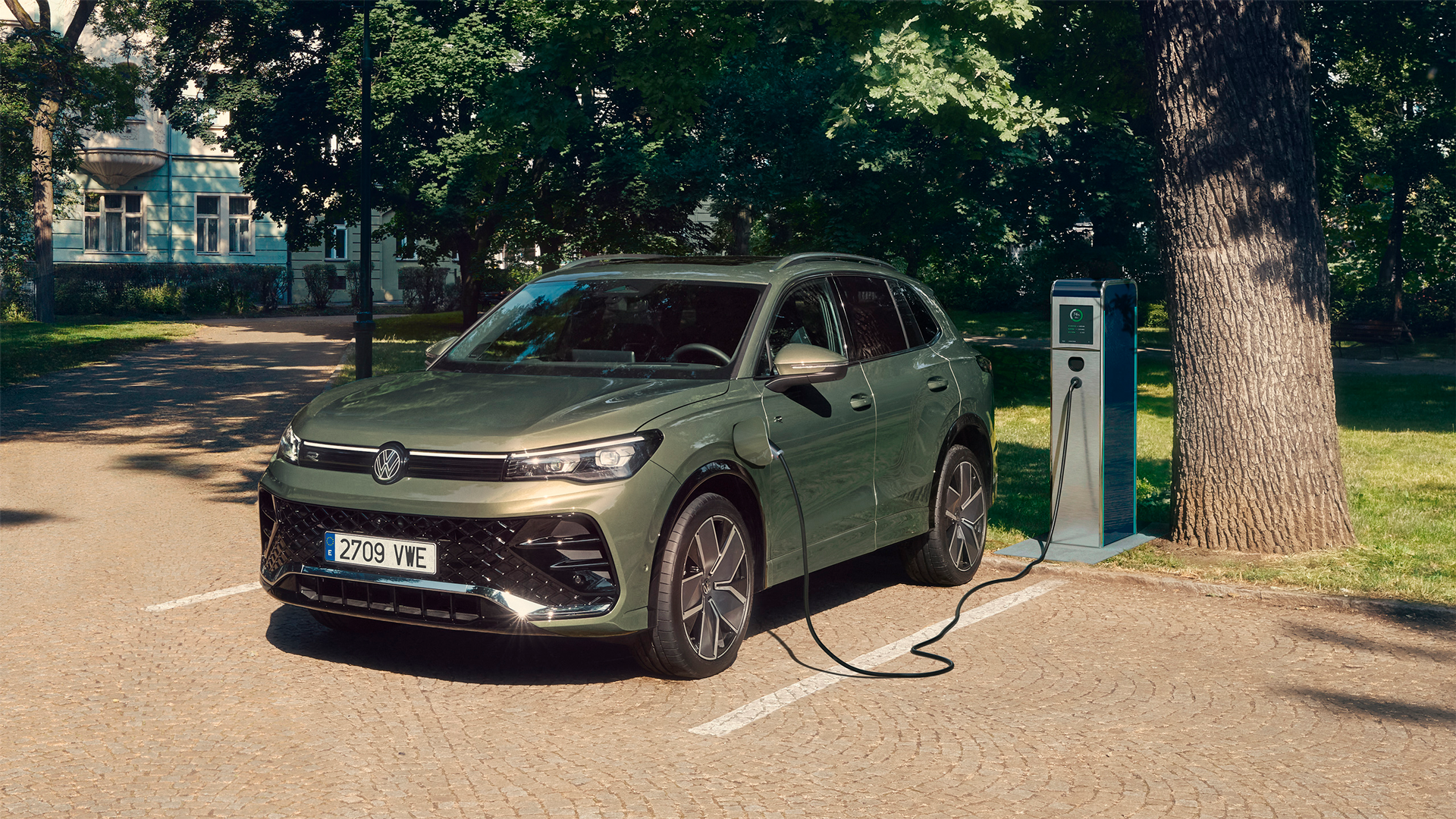 Cotxe Volkswagen verd oliva aparcat a l'exterior i connectat mitjançant un cable a una estació de càrrega elèctrica al costat d'uns arbres.