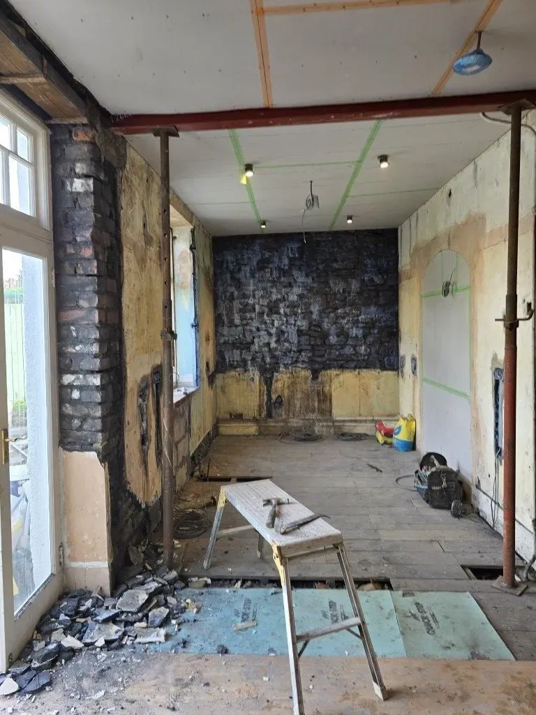 Unfinished lath and plaster walls and ceiling render first coat in a period property renovation in Redland, Bristol BS6. Visible are exposed floorboards, temporary site lighting with a yellow tripod work light, string lights, and electrical wiring. Preparation for a building conversion or new kitchen-diner space by JNS Plastering Services, showing initial render application and floor void work.