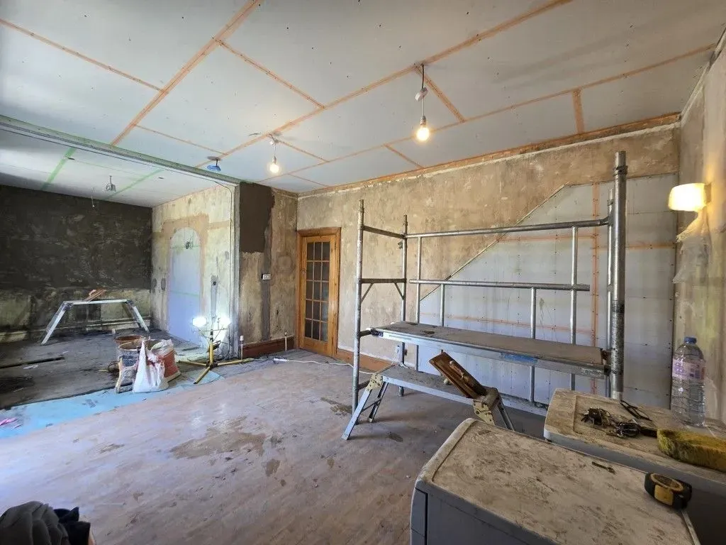 A view of an interior room undergoing a comprehensive renovation. The taped plasterboard ceiling is incomplete, the walls are partially stripped to show stone and old plaster, with temporary lighting. The floor void has blue sheeting, and open white French doors lead outside. A metal scaffolding tower and cement mixer are visible in the foreground, with bare concrete and wood floorboards.