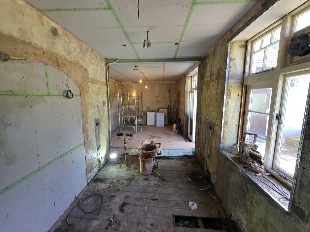 A view of an interior room undergoing a comprehensive renovation. The taped plasterboard ceiling is incomplete, the walls are partially stripped to show stone and old plaster, with temporary lighting. The floor void has blue sheeting, and open white French doors lead outside. A metal scaffolding tower and cement mixer are visible in the foreground, with bare concrete and wood floorboards.