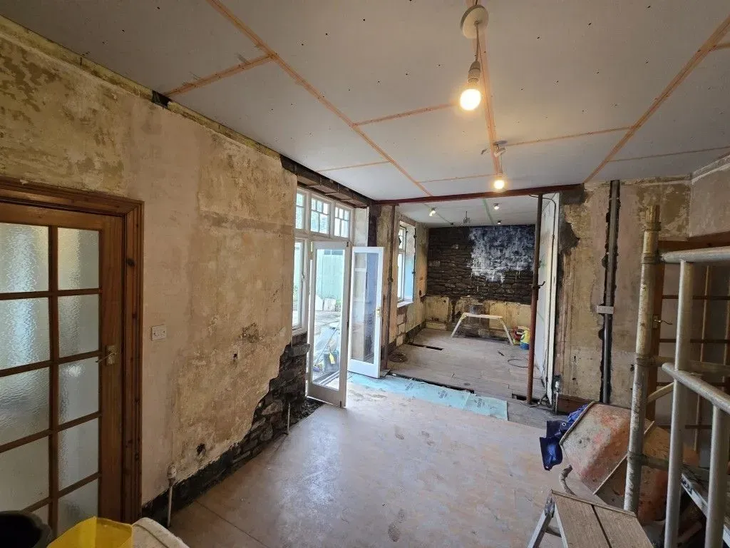 A view of an interior room undergoing a comprehensive renovation. The taped plasterboard ceiling is incomplete, the walls are partially stripped to show stone and old plaster, with temporary lighting. The floor void has blue sheeting, and open white French doors lead outside. A metal scaffolding tower and cement mixer are visible in the foreground, with bare concrete and wood floorboards.