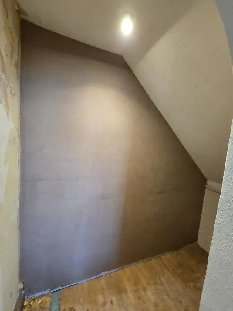 A shot of a small alcove or under-stairs space with a slanted ceiling, completely covered in smooth, dark plaster under a single glowing light fixture