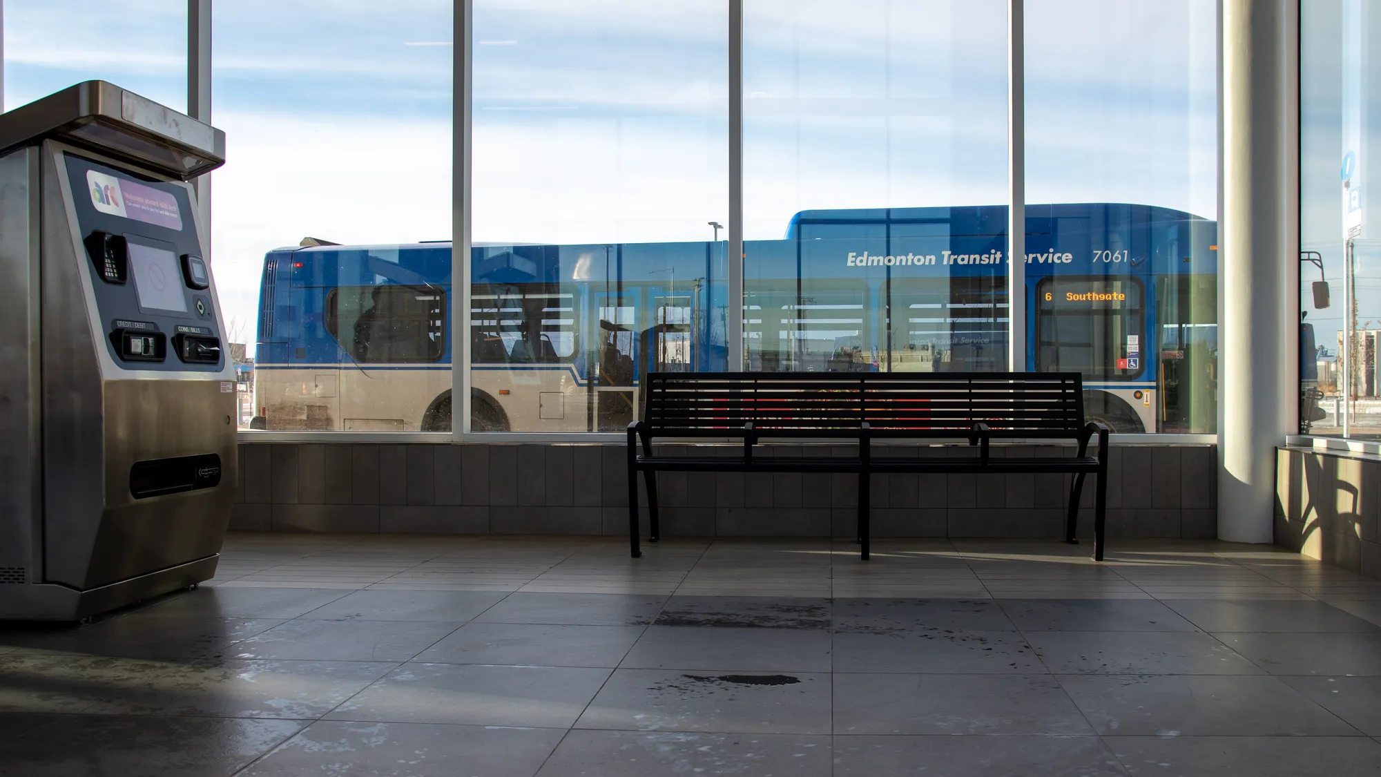 A black Modena bench sits inside a transit station, providing seating for passengers. The station is modern with large windows overlooking the bus terminal.