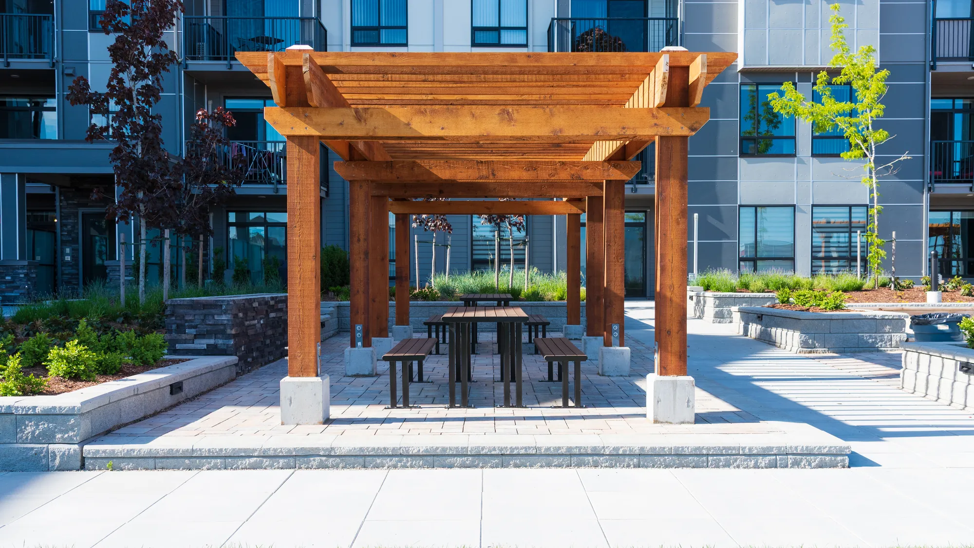 Outdoor picnic tables with benches provide a communal gathering space in a residential complex. The wooden pergola offers shade and shelter for residents to enjoy outdoor meals and socializing.