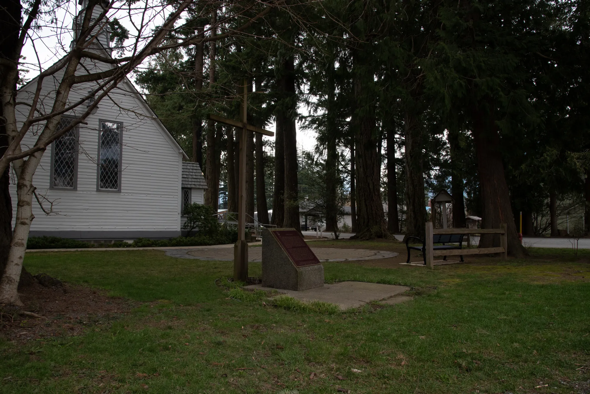 A Mountain Classic bench sits near a historic church in Hope, British Columbia. The bench provides a place to rest near a memorial and amidst tall trees.