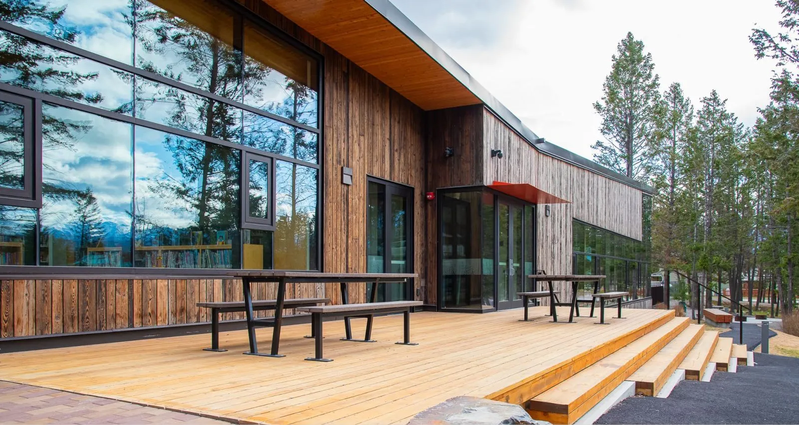 Outdoor picnic tables with benches provide seating outside a modern building with large windows. The tables feature a black frame and natural-toned lumber.