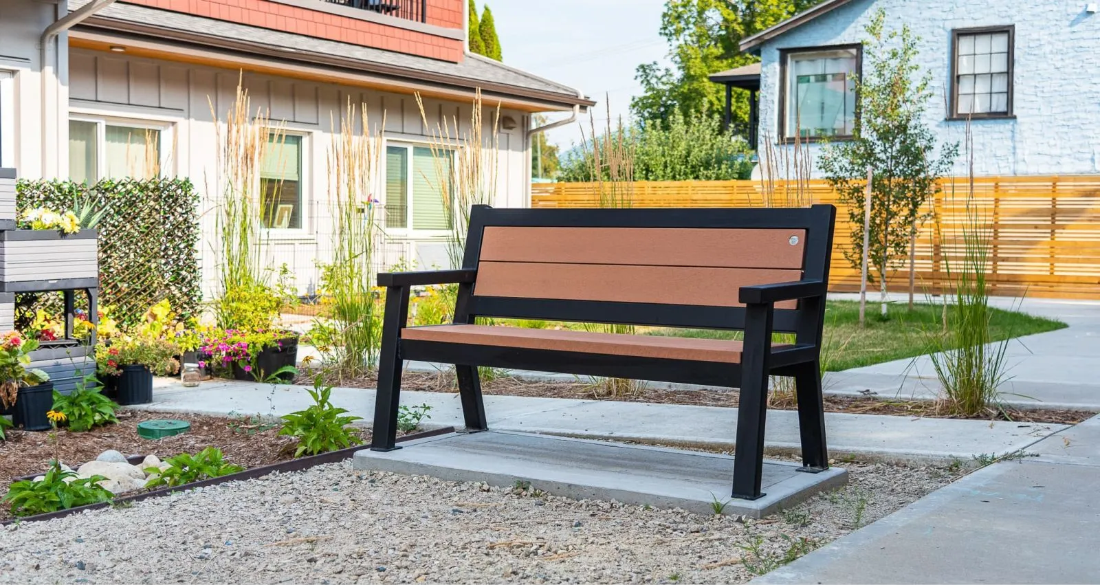 A Rutherford Angled Leg Bench provides seating in an outdoor setting with landscaping and nearby buildings. The bench features recycled plastic lumber and a powder-coated frame.