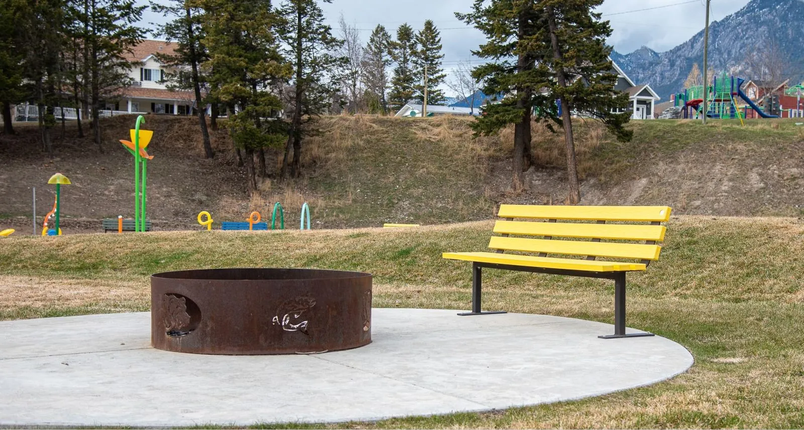 A yellow Standard Bench sits on a concrete pad in a park setting, providing a place to rest near a fire pit and playground. The bench offers a bright pop of color against the natural landscape.