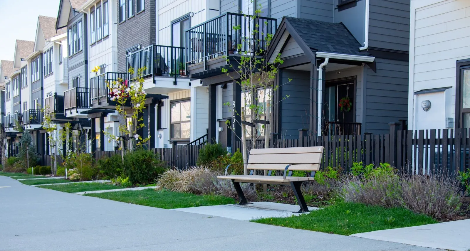 A Bayview bench provides seating along a sidewalk in a modern residential area. The bench features a light-colored recycled plastic lumber seat and black powder-coated metal frame.