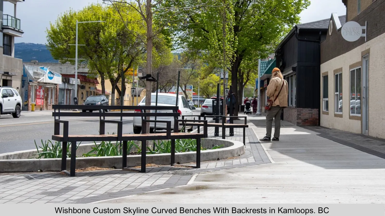 Curved benches with backrests line a sidewalk in Kamloops, British Columbia. The benches feature black frames and wood-look recycled plastic lumber.