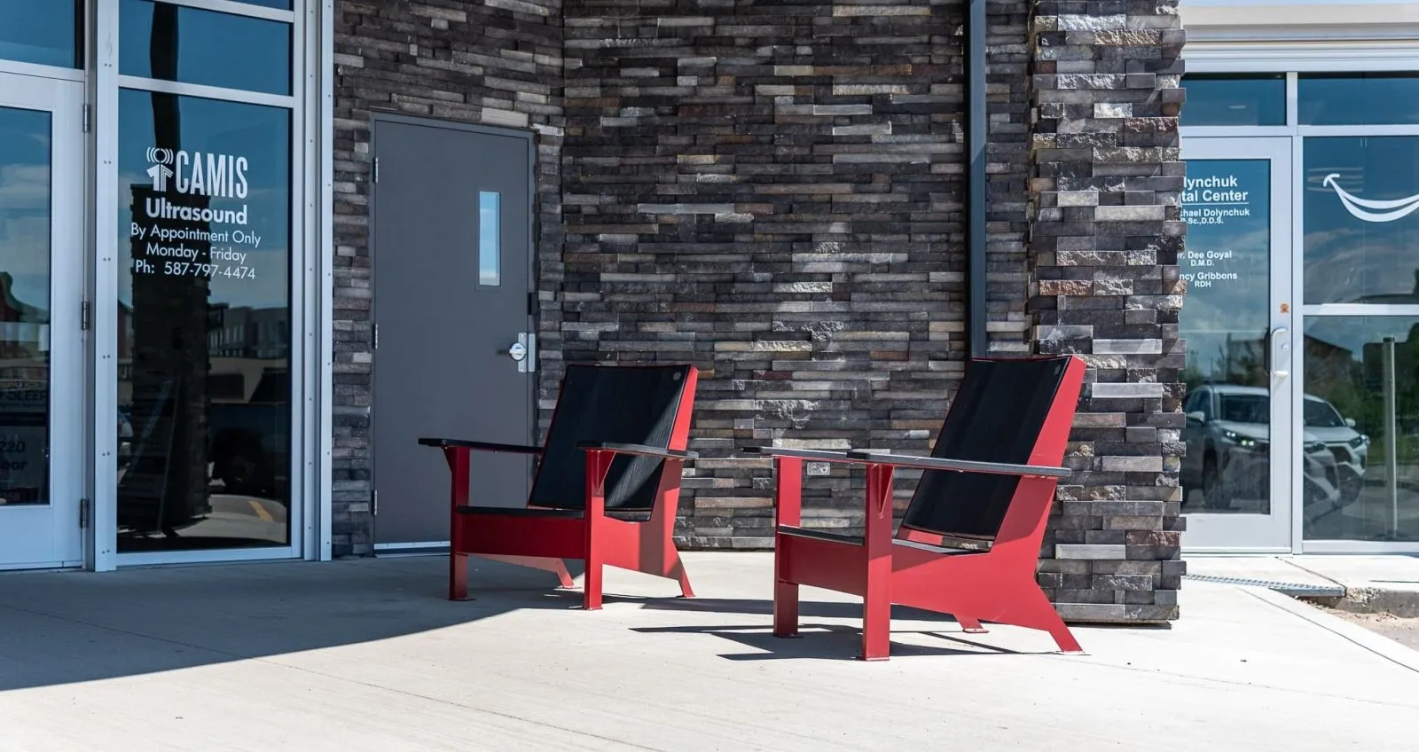 Two JEM Lounge Chairs provide seating outside a commercial building. The chairs feature a red frame and black seat.