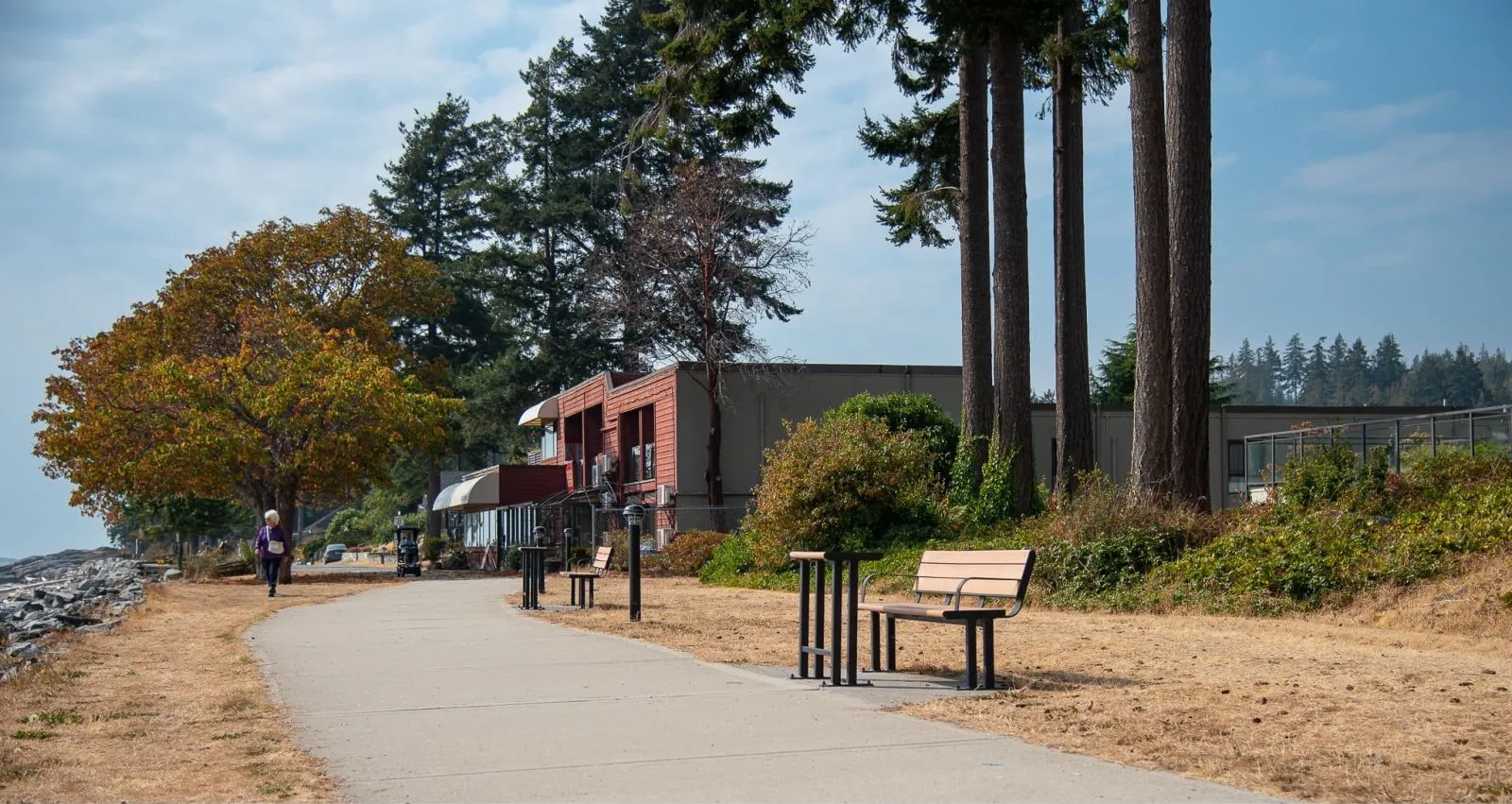 Larson benches line a paved path along the waterfront in Sechelt, British Columbia. The benches feature a simple design with vertical supports and horizontal slats.